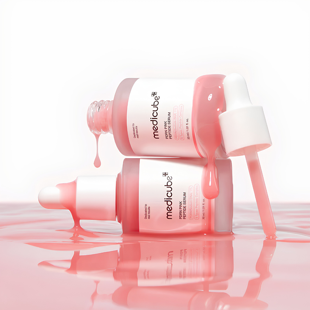 Two pink Medcube Hyaluronic Serum dropper bottles stacked and lying on their sides, with pink serum dripping from the bottles and a white dropper cap resting beside them, set against a clean white background with a pink reflective surface below.